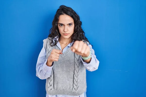 Young brunette woman standing over blue background punching fist to fight, aggressive and angry attack, threat and violence