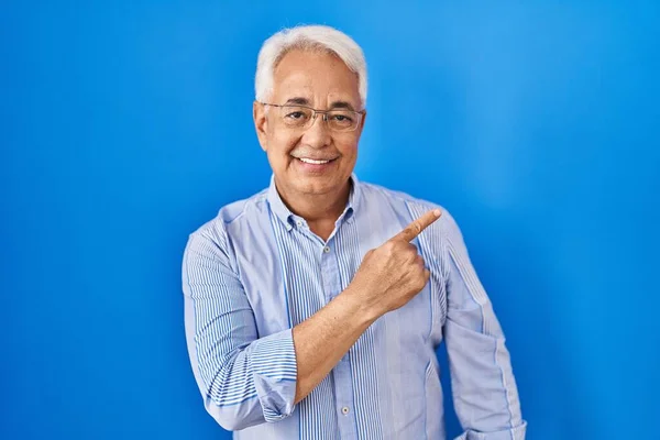 Hispanic senior man wearing glasses cheerful with a smile on face pointing with hand and finger up to the side with happy and natural expression