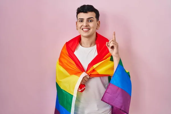 Non binary person holding rainbow lgbtq flag surprised with an idea or question pointing finger with happy face, number one