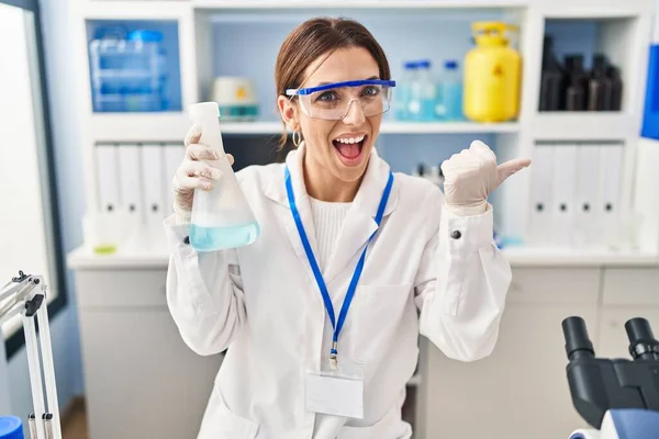 Young brunette woman working at scientist laboratory pointing thumb up to the side smiling happy with open mouth