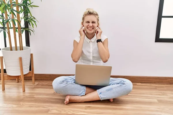 Young blonde woman using computer laptop sitting on the floor at the living room covering ears with fingers with annoyed expression for the noise of loud music. deaf concept.