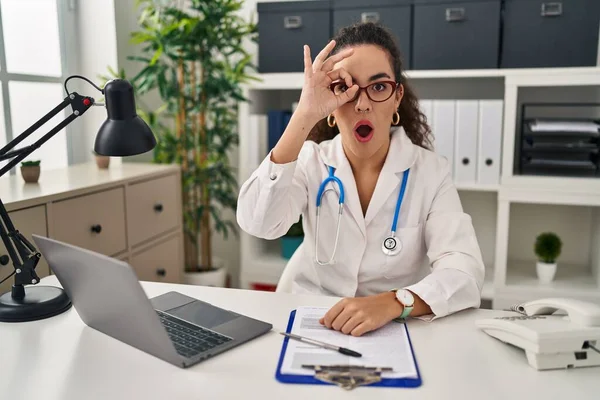 Young hispanic woman wearing doctor uniform and stethoscope doing ok gesture shocked with surprised face, eye looking through fingers. unbelieving expression.