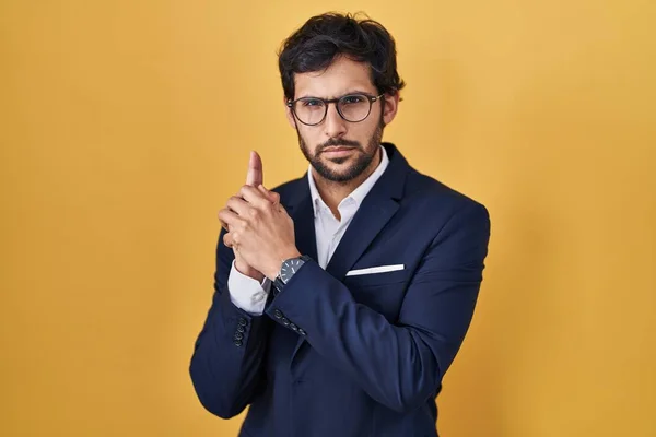Handsome latin man standing over yellow background holding symbolic gun with hand gesture, playing killing shooting weapons, angry face