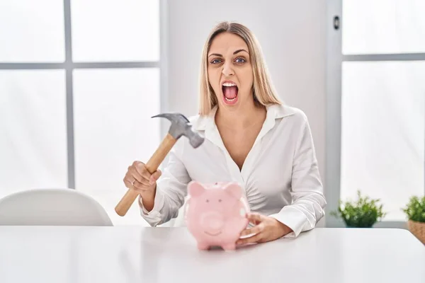 Young Blonde Woman Holding Piggy Bank Hammer Angry Mad Screaming — Stockfoto