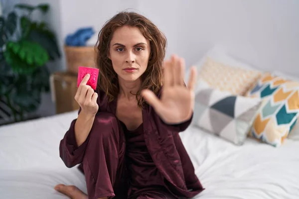 Beautiful brunette woman holding condom at the bedroom with open hand doing stop sign with serious and confident expression, defense gesture