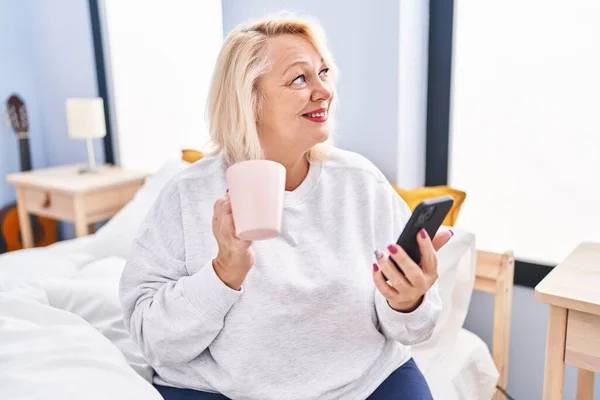 Middle age blonde woman using smartphone drinking coffee sitting on bed at bedroom