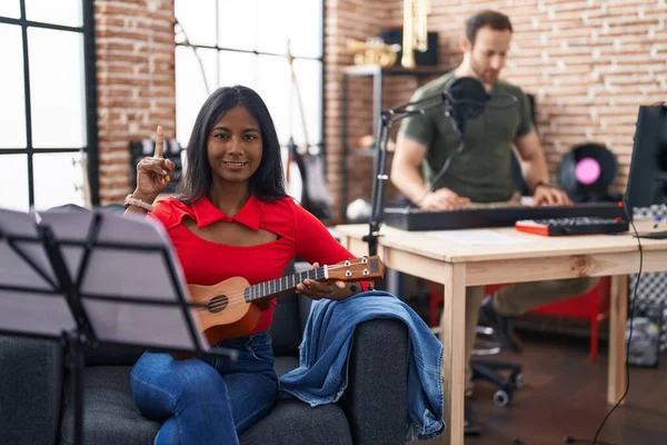 Young indian woman playing ukulele at music studio surprised with an idea or question pointing finger with happy face, number one