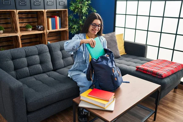 Young hispanic woman student holding books of backpack sitting on sofa at home