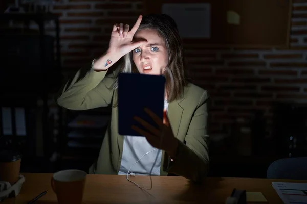 Blonde caucasian woman working at the office at night making fun of people with fingers on forehead doing loser gesture mocking and insulting.