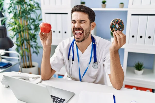 Mladý Hispánský Dietolog Držící Koblihu Jablko Rozzlobený Šílený Jekot Frustrovaný — Stock fotografie