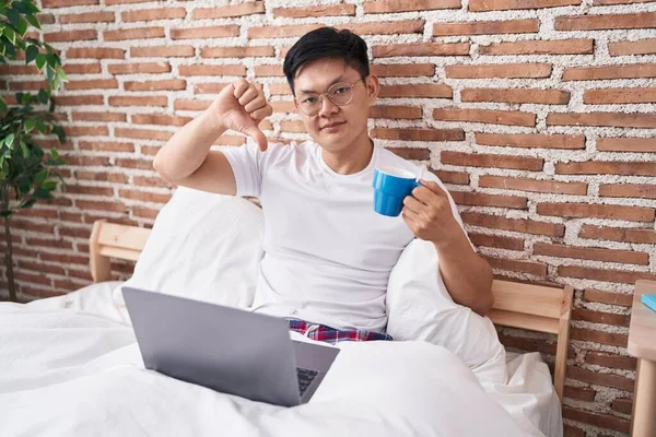 Young asian man drinking coffee sitting on the bed with angry face, negative sign showing dislike with thumbs down, rejection concept