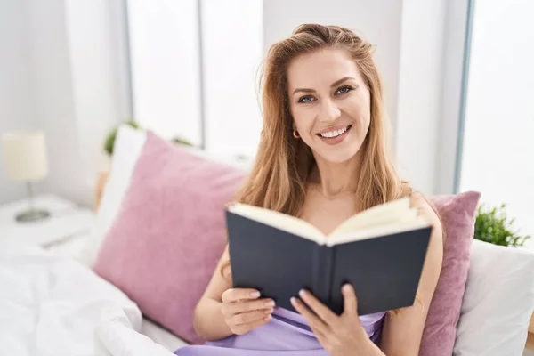 Young blonde woman reading book sitting on bed at bedroom