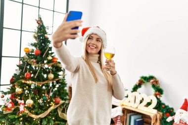 Genç sarışın kadın akıllı telefonun yanında selfie çekiyor. Noel ağacının yanında duruyor.
