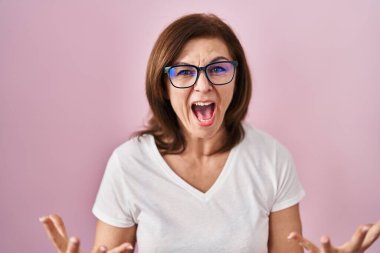 Orta yaşlı İspanyol kadın pembe arka planda deli deli deli bağırıyor ve agresif bir ifadeyle bağırıyor ve kollarını kaldırıyor. hüsran kavramı. 