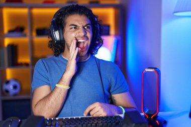 Hispanic man with curly hair playing video games clueless and confused with open arms, no idea and doubtful face. 