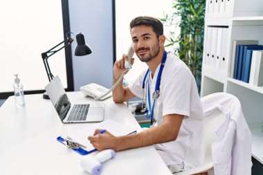 Young hispanic man wearing doctor uniform talking on the telephone working at clinic