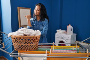 Young asian woman hanging clothes at clothesline smiling with happy face looking and pointing to the side with thumb up. 