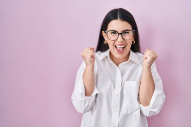 Young brunette woman standing over pink background celebrating surprised and amazed for success with arms raised and open eyes. winner concept. 