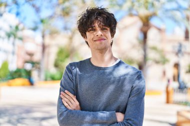 Genç İspanyol adam gülümsüyor. Parkta kollarını kavuşturmuş duruyor.