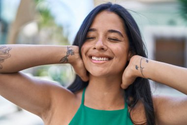 Young hispanic woman smiling confident relaxed with hands on head at street