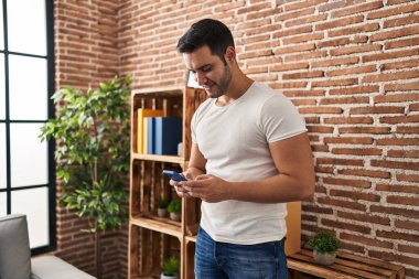 Genç İspanyol adam evde akıllı telefon kullanarak kendinden emin gülümsüyor.