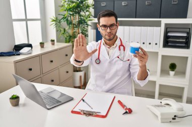 Young hispanic doctor man with beard holding ear cotton buds with open hand doing stop sign with serious and confident expression, defense gesture 