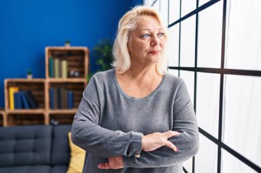 Orta yaşlı sarışın kadın evde kollarını kavuşturup el kol hareketi yapıyor.