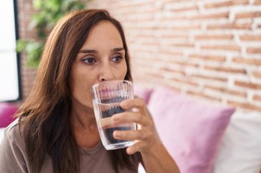 Young beautiful hispanic woman drinking glass of water sitting on bed at bedroom