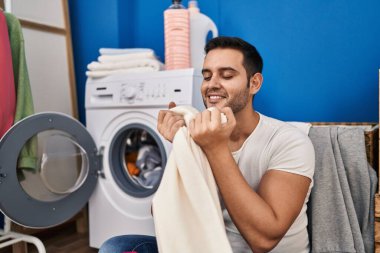 Young hispanic man smiling confident smelling clothes at laundry room