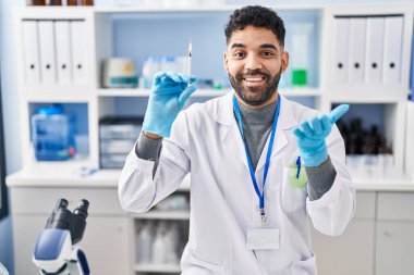 Hispanic man with beard working at scientist laboratory holding syringe celebrating achievement with happy smile and winner expression with raised hand 