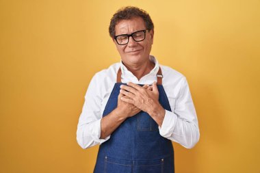 Middle age hispanic man wearing professional cook apron smiling with hands on chest with closed eyes and grateful gesture on face. health concept. 