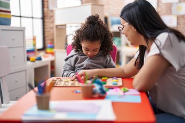 Teacher and toddler playing with maths puzzle game sitting on table at kindergarten