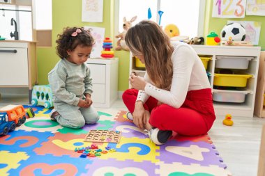 Teacher and toddler playing with maths puzzle game sitting on floor at kindergarten