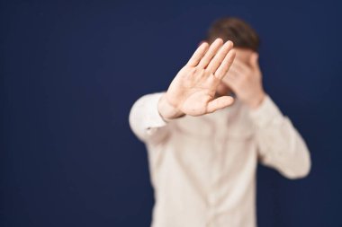 Mavi arka planda sakallı İspanyol bir adam elleriyle gözlerini kapatıyor ve üzgün ve korku ifadesiyle dur hareketi yapıyor. utangaç ve olumsuz kavram. 