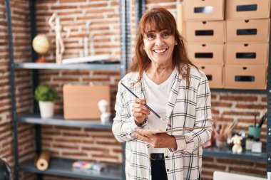 Middle age woman artist smiling confident holding paintbrush and palette at art studio