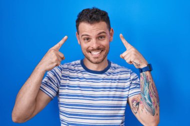 Mavi arka planda duran genç İspanyol adam gülümsüyor iki eliyle kafasını işaret ediyor, harika bir fikir ya da düşünce, iyi bir hafıza 