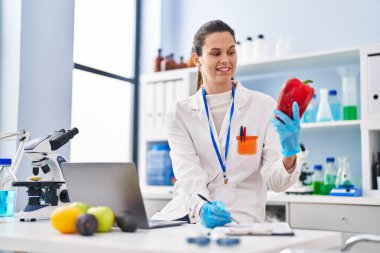 Young beautiful hispanic woman scientist holding pepper writing on document at laboratory
