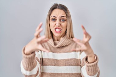 Young blonde woman wearing turtleneck sweater over isolated background shouting frustrated with rage, hands trying to strangle, yelling mad 