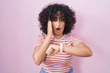 Young middle east woman standing over pink background looking at the watch time worried, afraid of getting late 