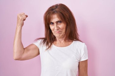 Middle age woman standing over pink background strong person showing arm muscle, confident and proud of power 