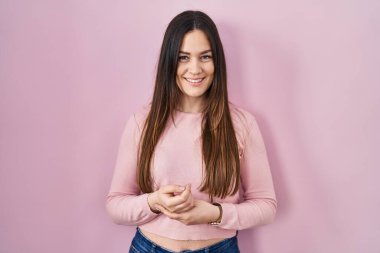 Young brunette woman standing over pink background with hands together and crossed fingers smiling relaxed and cheerful. success and optimistic 