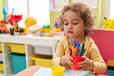 Adorable hispanic toddler student sitting on table drawing on paper at kindergarten