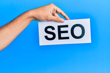 Hand of caucasian man holding paper with seo word over isolated blue background