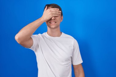 Caucasian blond man standing over blue background smiling and laughing with hand on face covering eyes for surprise. blind concept. 