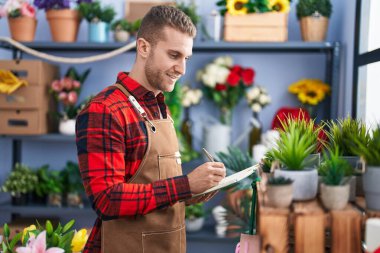 Young caucasian man florist smiling confident writing on notebook at flower shop