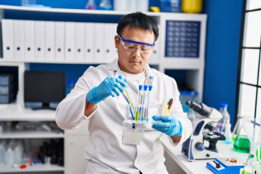 Young chinese man wearing scientist uniform holding test tubes at laboratory