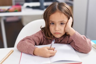 Adorable hispanic girl student writing on notebook with relaxed expression at classroom