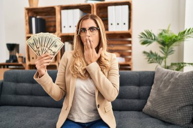 Young blonde therapist woman working at therapy office holding money covering mouth with hand, shocked and afraid for mistake. surprised expression 
