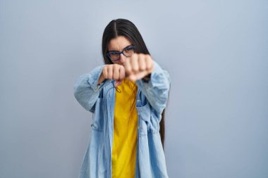 Young hispanic woman standing over blue background punching fist to fight, aggressive and angry attack, threat and violence 