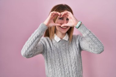Beautiful woman standing over pink background doing heart shape with hand and fingers smiling looking through sign 